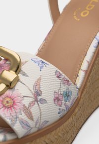 Hvid kile-sandal med blomsterprint, guldlåsdetalje og flettet sål i strå.