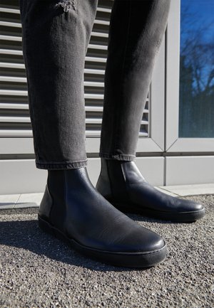 Persona che indossa stivaletti in pelle nera e jeans neri strappati, in piedi sull'asfalto vicino a un edificio con rivestimento in metallo.