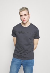 Juodas medvilninis marškinėliai su apvalia iškirpte, su toniniu spausdintu logotipu "ALPHA INDUSTRIES" ir grafikos akcentais priekyje.