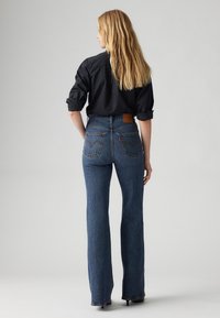 High-waisted denim flared jeans in a dark blue wash. Features classic back pockets with stitching detail and a leather patch at the waistband.