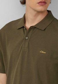 Olivgrünes Polohemd aus strukturiertem Baumwollstoff. Verfügt über einen Kragen, zwei Knöpfe und ein goldenes "s.Oliver"-Logo auf der linken Seite.
