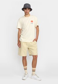 Camiseta crema con gráfico de sol naranja y texto teal, combinada con pantalones cortos amarillos claros, sombrero bucket gris, zapatillas blancas y calcetines a rayas.