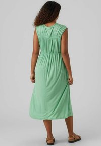 Vestido verde menta hasta la rodilla con mangas tipo capa, detalle de fruncido en la espalda y cintura elástica. La tela parece suave y lisa.