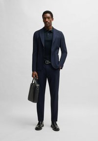 Marinblå blazer med texturerad yta, matchad med en mörk polo och byxor. Tillbehör inkluderar en svart läderväska. Svarta dressade skor.