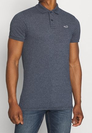 Man draagt een nauwsluitend donkergrijs poloshirt met korte mouwen, drie knopen en een klein wit logo op de borst.