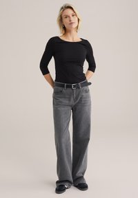 Un haut noir à manches trois-quarts associé à un jean gris taille haute à jambes larges et une ceinture noire, le tout sur un fond neutre.