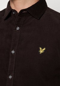 Camisa de pana marrón con cuello abotonado, que presenta un pequeño logo de águila bordado en amarillo y botones grises brillantes.