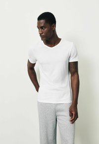 Fehér pamut rövid ujjú V-nyakú póló, testhezálló szabás, sima textúra, szürke melegítőnadrággal párosítva. A modell kezeit a csípőjén pihenteti.