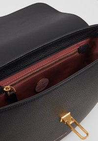 Borsa a mano in pelle nera texturizzata con chiusura dorata, parzialmente aperta per mostrare l'interno in tessuto color bordeaux e la tasca con cerniera con etichetta del marchio.