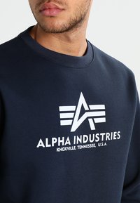 Sweatshirt bleu marine avec un logo blanc et le texte "ALPHA INDUSTRIES" sur le devant. Design à encolure ronde avec des poignets et un ourlet côtelés.