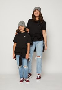 Två individer bär svart t-shirt med en vit logga, i kombination med slitna ljusblå jeans och gråa mössor, stående mot en enkel bakgrund.