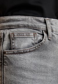 Acercamiento de un bolsillo de jeans de mezclilla gris claro con costuras marrones visibles y una camiseta negra metida en la cintura.