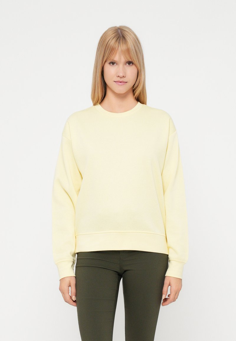 Geltona palaidinė laisva fasonu, su ilgomis rankovėmis, džinsiniu apykakliniu kraštu ir švelnia tekstūra, derinama su tamsiai žaliomis prigludusiomis kelnėmis.