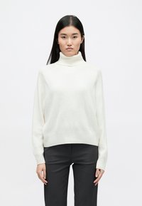 Λευκό πουλόβερ με ψηλό λαιμό και ραβδωτή υφή, φαρδιά εφαρμογή και χαμηλούς ώμους, συνδυασμένο με σκούρες παντελόνες. Απλός σχεδιασμός, χωρίς ορατά μοτίβα.