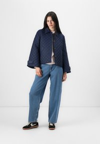 En ung kvinne står mot en hvit bakgrunn, iført en marineblå quiltet jakke over en lys genser og vide jeans, kombinert med svarte sneakers.