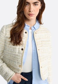 Giacca in tweed crema con un motivo strutturato, fili dorati, bottoni rotondi e un design cropped, indossata sopra una camicia blu chiaro.