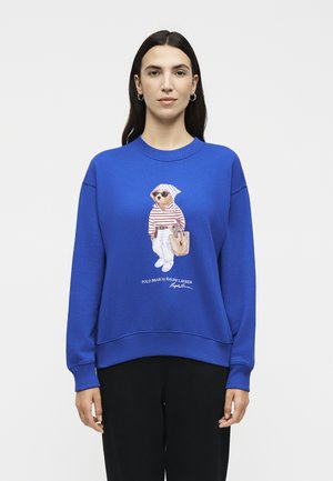 Γυναίκα με μακριά σκούρα μαλλιά που φορεί ένα μπλε φούτερ με μια αρκούδα με γυαλιά ηλίου, ριγέ πουκάμισο, λευκό παντελόνι και κρατά μια τσάντα tote.