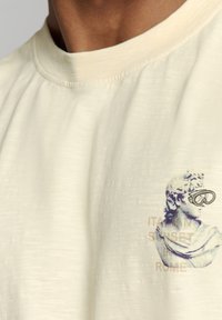 Crème-kleurige katoenen t-shirt met een textuurafwerking en een graphic van een klassieke standbeeld met een bril, met de tekst "ITALIAANSE ZONSONDERGANG ROME".