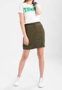 Jupe vert olive avec taille à cordon et poches latérales, assortie à un t-shirt blanc avec des lettres vertes. Baskets blanches.