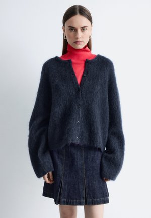 Femme aux cheveux raides portant des boucles d'oreilles argentées en forme d'anneau, un col roulé rouge, un cardigan bleu foncé duveteux et une jupe plissée en denim foncé, face à l'avant.