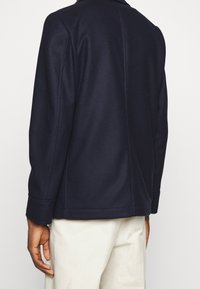 Chaqueta azul marino con una textura suave, corte ajustado y detalles de costura visibles en la parte posterior. Mangas largas con puños.