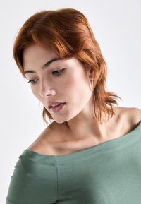 Grønn off-shoulder topp med myk, lett stoff; modell med kort kastanjefarget hår iført en enkel hoop-øredobb mot en lys bakgrunn.