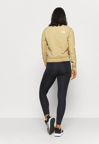 Beige mikina s logem na zadní straně, spárovaná s černými legínami s texturovanými detaily a černými sportovními botami s bílou podrážkou.