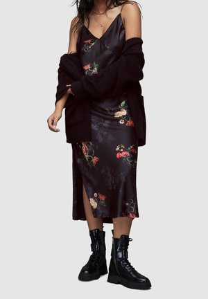 Femme portant une robe nuisette noire à motifs floraux avec une fente latérale, un cardigan noir posé sur les épaules et des bottes de combat noires sur fond gris.