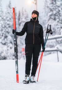 Langläufer in schwarzer isolierter Jacke und Hose, hält orangefarbene Skier und Stöcke, trägt Schuhe mit blauen Akzenten vor einem verschneiten Hintergrund.