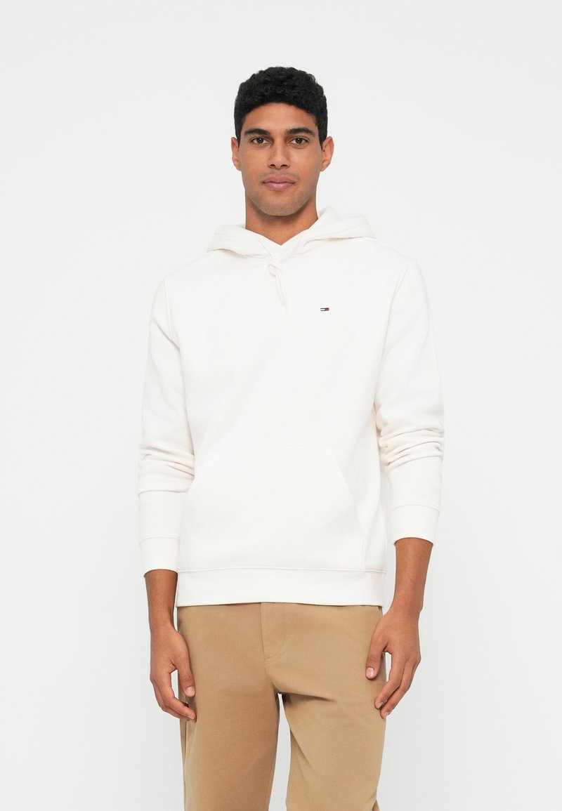 Muž oblečený v bielej mikine s kapucňou a predným vreckom, so malým logom, v kombinácii s béžovými nohavicami, stojí na pozadí v bielej farbe.