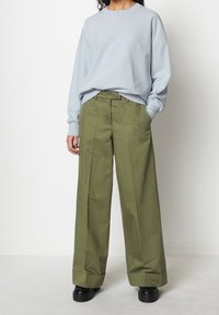 Persona che indossa un oversized maglione azzurro chiaro, pantaloni larghi verde oliva con risvolti e scarpe con piattaforma nere, in piedi contro un muro bianco.