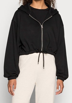 Mujer que lleva una sudadera con capucha negra corta con cremallera, cordón en la cintura y mangas largas, combinada con pantalones de tiro alto en beige claro.