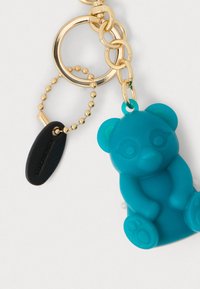 Μπρελόκ αρκουδάκι από σιλικόνη σε τιρκουάζ χρώμα, με χρυσή αλυσίδα και διακοσμητικό χάντρα, καθώς και μαύρη ετικέτα με την ένδειξη 'Κατασκευάστηκε στην Κίνα'. Ομαλή υφή σε όλο το μπρελόκ.