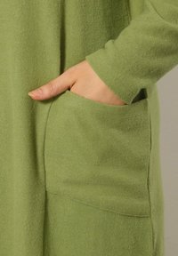 Groene zachte stof met een zijzak en lange mouwen. De textuur lijkt gezellig en licht opgeruwd, met een gestroomlijnde pasvorm.