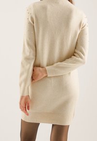 Robe pull tricotée de couleur crème avec de longues manches, poignets et ourlet côtelés, ornée de petites perles décoratives sur les épaules. Texture lisse.