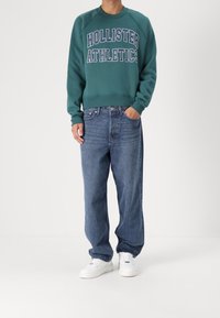 Felpa verde con la scritta bianca "Hollister Athletics", abbinata a jeans blu larghi in denim e sneaker bianche; vestibilità comoda e design casual.