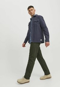 Navy blue button-up jacket with chest pockets, layered over a light blue shirt, paired with olive green pants and beige shoes.