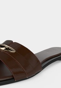 Sandale slide marron avec deux larges brides et une boucle ovale dorée sur une semelle plate noire avec une semelle intérieure de marque.