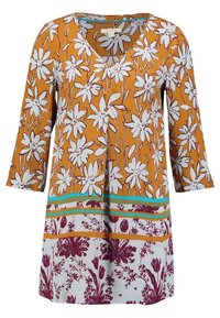 Tunique florale en jaune moutarde avec des fleurs blanches. Elle présente des manches 3/4, un décolleté en V, et des détails de motifs superposés en teal et bordeaux au bord.