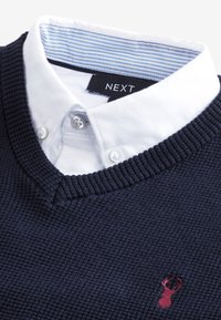 Marineblå struktureret sweater med V-hals, lagt over en hvid skjorte med krave. Har en blå stribet krave og et lille rødt hjortelogo.