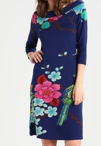 Marinblå klänning med trekvartslånga ärmar, med stora blommönster i rosa, grönt och ljusblått, med en fågeldekoration.