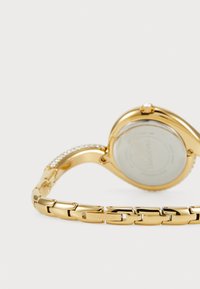 Kuldne metallkell sileda korpuse, bändi kriystalaktsentide ja läbipaistva tagaküljega, kus on nähtav brändi nimi "Swarovski".