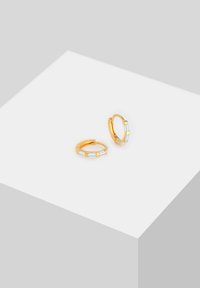 Boucles d'oreilles créoles dorées mettant en valeur des pierres claires rectangulaires, avec une texture lisse et un design minimaliste, posées sur une surface blanche.