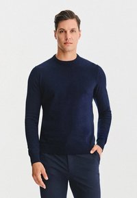 Granatowy sweter z okrągłym dekoltem, wykonany z miękkiej, teksturowanej tkaniny. Dopasowany krój z długimi rękawami i ściągaczami przy mankietach. Połączony z ciemnymi spodniami.