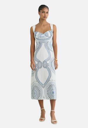 Kobieta z zaplecionymi włosami, ubrana w białą sukienkę midi z niebieskimi okrągłymi wzorami, białe buty na obcasie z paskiem wokół kostki i złotą bransoletkę.