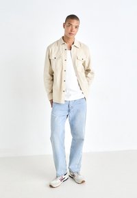 Camicia button-up beige con due tasche sul petto sopra una maglietta bianca, abbinata a jeans azzurri chiari e sneakers grigie con accenti rossi e blu.
