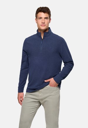 Modelo masculino vistiendo un pulóver azul marino con cremallera hasta el pecho y pantalones beige claros, de pie con una mano en el bolsillo sobre un fondo liso.