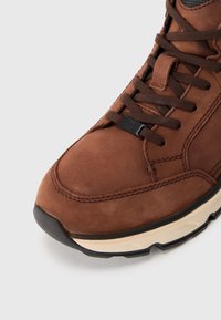 Sneaker in pelle marrone con superficie texture, punta rotonda, suola in gomma nera e lacci marroni. Presenta un piccolo logo sul lato.