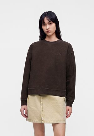 Donna con felpa marrone scuro a maniche lunghe e gonna di jeans beige chiaro, in piedi davanti a uno sfondo bianco semplice.