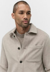 Beige ullblandning jacka med krage, knappstängning och framficka. Under jackan en texturerad stickad tröja i en ljusare nyans.